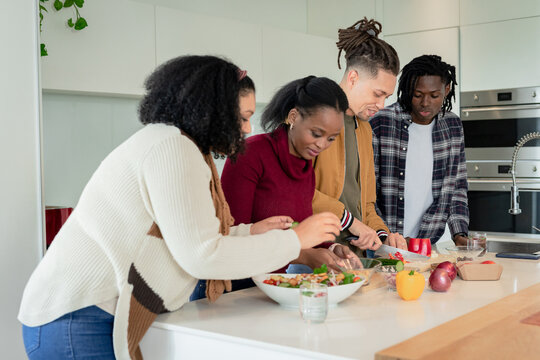 Diverse friends mixing salad in bowl and chopping vegetables with chef's knife on kitchen island