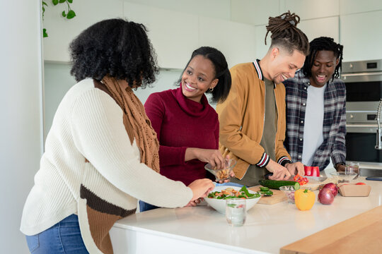 Diverse friends chopping vegetables, tossing salad in glass bowls on white island in modern kitchen