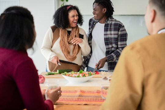 Diverse friends laughing and slicing vegetables into white salad bowl at modern kitchen island