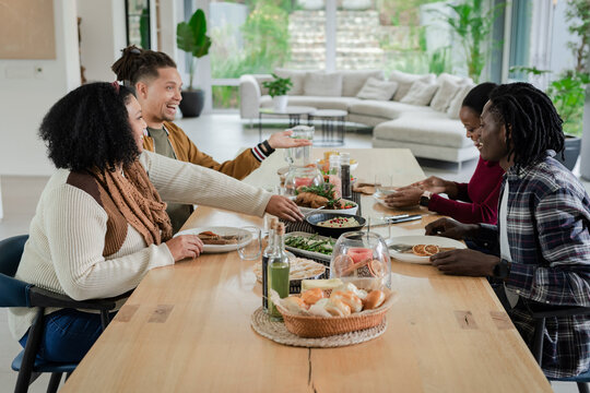 Diverse friends sharing meal around wooden table at home with basket of rolls and salad - Powered by Adobe