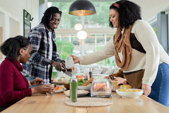 Diverse friends gathering at dining table sharing meal pouring from white pitcher and passing salad