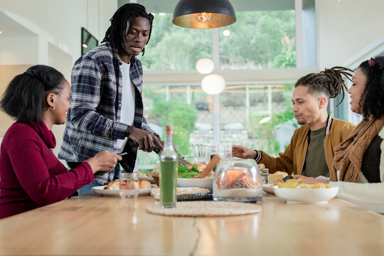 Diverse friends serving salad and cutting bread at dining table with salad bowl, copy space