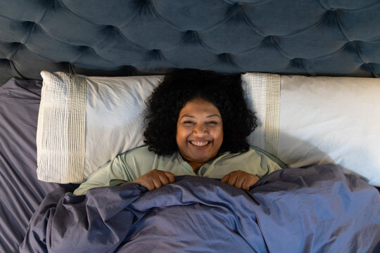 African American woman lying on bed wearing pajamas holding duvet near chest with tufted headboard - Powered by Adobe