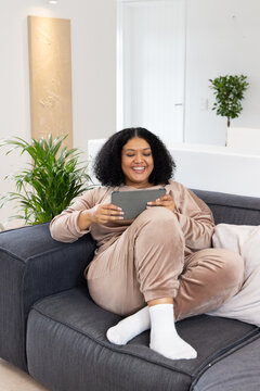 Mid adult African American woman wearing tracksuit sitting on couch in living room holding tablet