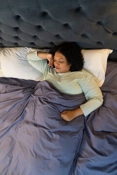 Woman lying in bedroom with tufted headboard, covered by gray duvet, wearing pale green top