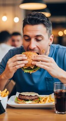 A man mid-bite into a hamburger. French fries and a drink are in front of him. A blurred restaurant setting in the background