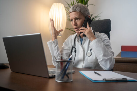 Mature female doctor stressed in clinic, looking at laptop while handling phone calls, worried and tense about work and mistakes.