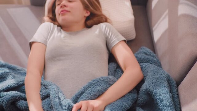 Sleepy girl finally wakes up stretching herself on couch holding soft blanket, eyes closed with relaxed expression as warm morning sunlight fills room