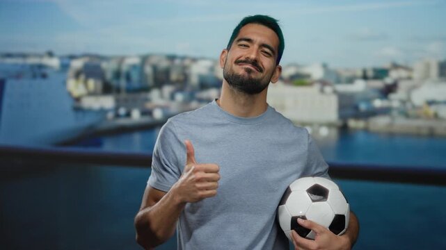 Young man smiling confidently by seaside holding soccer ball with thumbs up in casual shirt outdoors showcasing happiness and leisure with vibrant backdrop of port.
