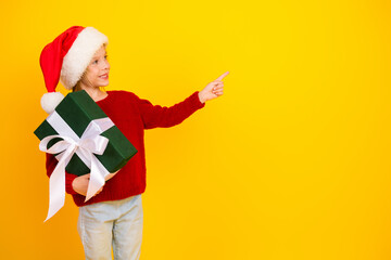 Playful child in Santa hat with a green gift points to the right against a bright yellow background...