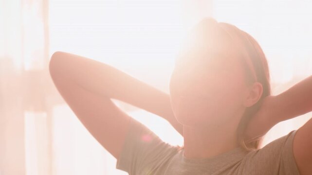 Close up of woman stretching muscles after waking up with gentle smile on face, arms raised behind head, morning sunlight streaming through window creating warm glowing peaceful atmosphere
