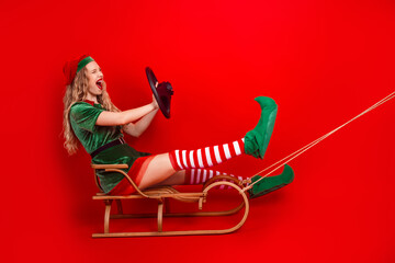 Festive elf in a cheerful pose riding a wooden sled against vibrant red backdrop delivers holiday...