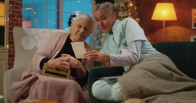 Caregiver and senior woman sit together at home during Christmas, enjoying old photos and stories, reliving the past with warmth, laughter, and heartfelt connection.