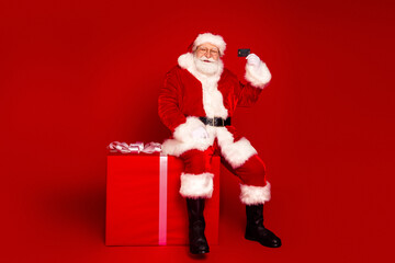 Santa Claus sits on a large gift box taking a selfie with a red background celebrating Christmas...