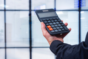 Hand in a business suit holding a black calculator against a blurred background of a modern office or corporate interior. Concept of financial management, accounting and business calculations.