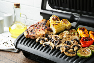 Electric grill with steak and vegetables on wooden table in kitchen, closeup