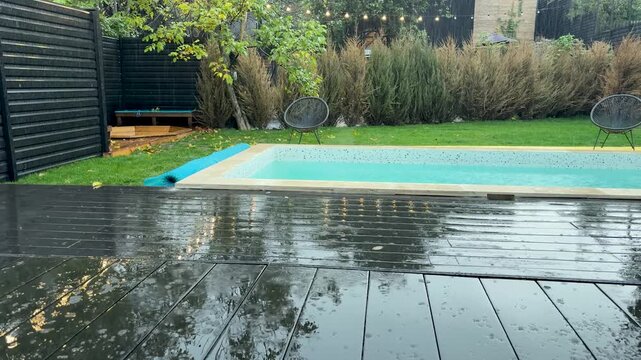 Heavy rain falling on the wet wooden deck of a modern backyard swimming pool during a gloomy autumn day, creating ripples on the water surface and reflecting the surrounding nature
