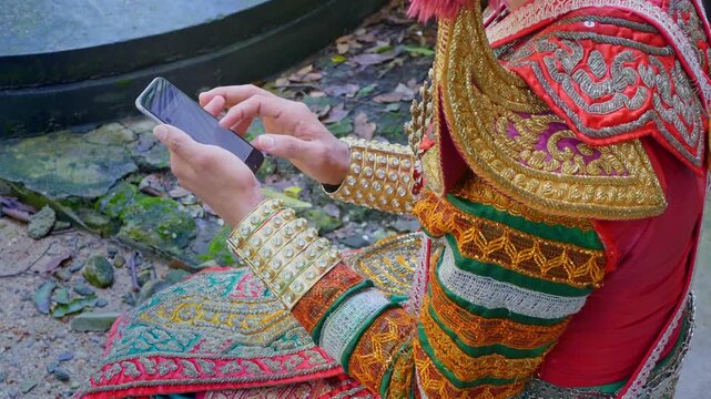 A Nora dancer dressed in vibrant traditional costume takes a quiet break, sitting gracefully while using her smartphone. The contrast between heritage and modernity captures Thailand&rsquo;s living culture.