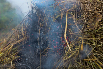 ​A close-up shot depicts a smoldering fire in a pile of dry grass or hay, with visible red embers and bluish smoke rising upwards. The image conveys the destructive power of fire and the process of bu