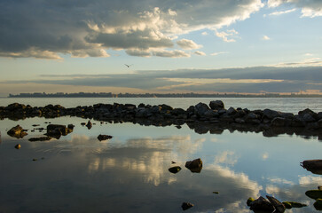 Naklejka premium Dramatic Sky Reflection on Water with Rocky Shore and Distant City Silhouette