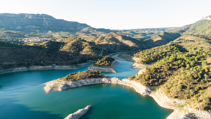 Obraz premium Aerial view of a mountain reservoir in Spain with turquoise water surrounded by forested hills, rocky shores, and a dam under clear skies