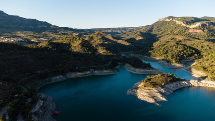 Aerial view of a mountain reservoir in Spain with turquoise water surrounded by forested hills, rocky shores, and a dam under clear skies