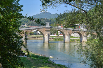 Fototapeta premium A historic stone bridge spans a calm river, surrounded by greenery and backed by forested hills. Konjic town on Neretva river, Bosnia and Herzegovina