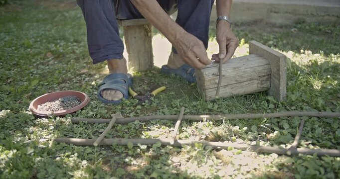 A skilled craftsman meticulously assembles a charming wooden ladder or trellis using natural sticks, a hammer, and nails in a vibrant garden setting on a sunny day.