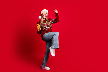 Fototapeta premium Playful young woman in santa hat jumps with smartphone in hand against a bold red background celebrating christmas