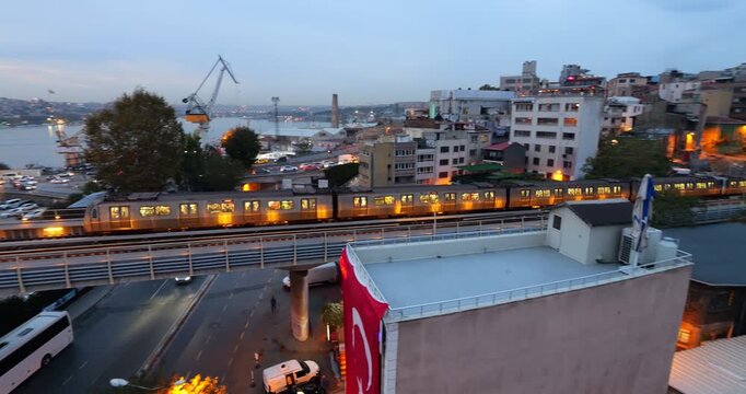 Evening view of metro train riding on viaduct above city road, approaching Golden Horn Metro Bridge in Istanbul. Warm twilight glow and tracking shot highlight train in motion against urban view