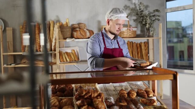 Man seller stands at bakery counter with basket filled with fresh croissants. Employee puts croissants in window, arranges display of goods, complements assortment, puts buns on sale 