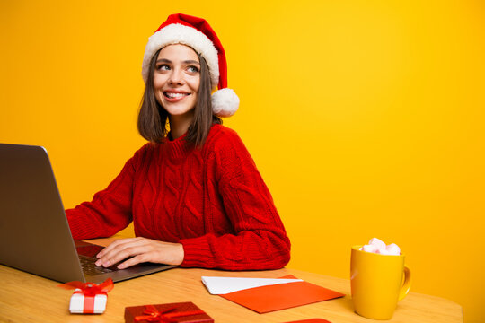 Funny festive office style woman in santa hat red sweater smiling at laptop in bright yellow studio mood