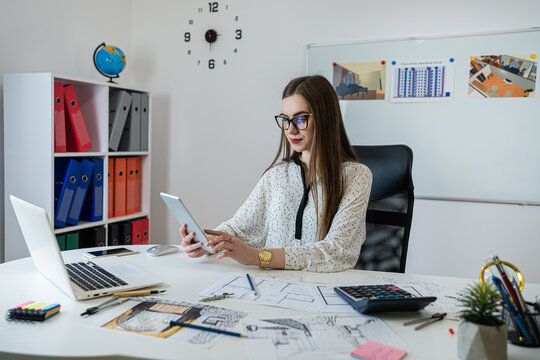 Pretty young woman working of architect blueprint at desk in modern office - Powered by Adobe