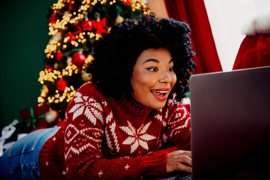 Joyful young woman uses a laptop while relaxing near a warmly decorated Christmas tree, embracing the festive spirit.