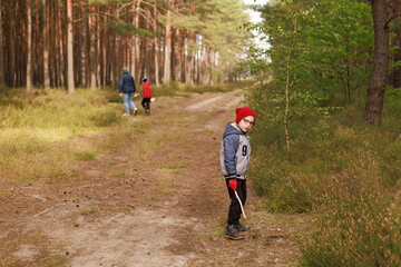 Obraz premium Boy in red hat stands on forest trail, with father and brother collecting mushrooms in the background. Concept of family leisure, nature exploration, and autumn atmosphere.