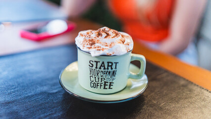 Happy young woman holding a rustic ceramic mug with a gourmet coffee drink topped with whipped cream and chocolate powder. The mug has the text "Start your morning with a cup of coffee". Ideal for con