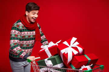 Playful man in a christmas sweater with scarf enjoys shopping in a cart full of gifts against a...