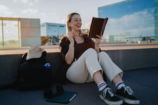 Young woman sitting outdoors with notebook and pen, smiling while writing, symbolizing creativity, education, digital nomad lifestyle and modern freelance inspiration. - Powered by Adobe