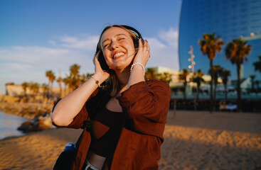 Cheerful young woman enjoying music with headphones, eyes closed, smiling wide under golden hour...