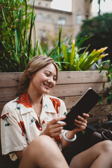 Asian young woman sitting comfortably, looking at tablet with pleasant smile, holding stylus in natural light, enjoying relaxed moment in peaceful freelance work setting. © BullRun
