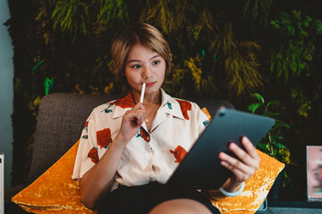 Asian woman with stylus in hand, staring at tablet thoughtfully, symbolizing online study, focus, and adaptability in digital freelance work. © BullRun