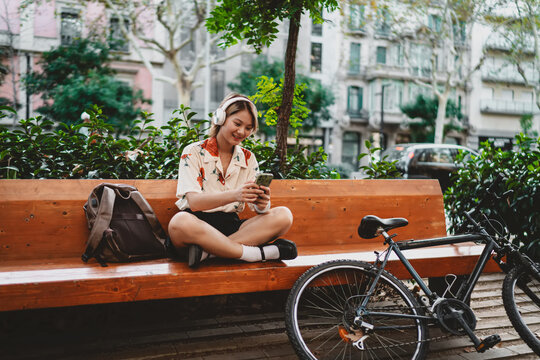 Asian woman smiles while sitting on bench with legs wide on bike, floral shirt and headphones on. Evokes carefree digital lifestyle, relaxation, and smart self-directed creativity outdoors.
