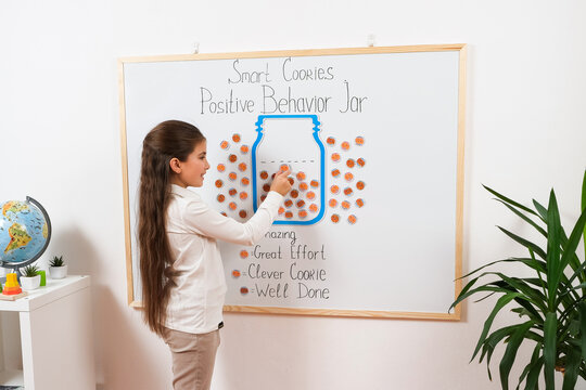 A young schoolgirl adds reward stickers to a positive behavior chart in a classroom. The educational activity promotes motivation, responsibility, and encouragement for good behavior.