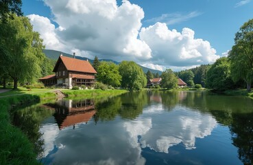 Obraz premium Traditional wooden houses near tranquil lake with reflections. Rich green trees and hills form scenic landscape. Peaceful nature with clouds in blue sky.