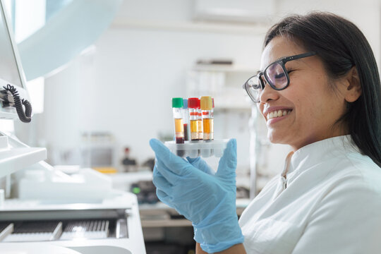 A lab technician smiles while examining blood samples in a modern laboratory setting during the day.