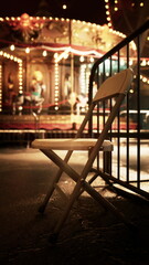 A wooden chair sits in front of an illuminated carousel in an abandoned amusement park at night.