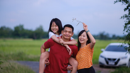 The daughter happily rides on her father's back with her smiling mother beside her.Happy family traveling on holiday by car. Parents and daughter spend free time together