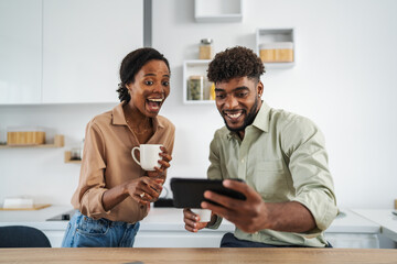 Happy black couple laughing watching video on phone in modern kitchen