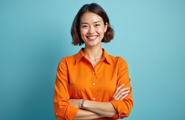 Attractive Asian woman in orange shirt smiles. Lady with short bob hair style poses against a blue backdrop. Smiling businesswoman with folded arms looks at camera. Office worker is in good mood