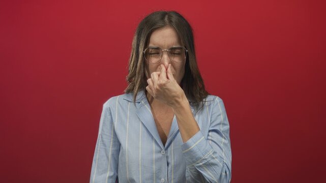 Young hispanic woman wearing glasses and striped shirt pinches nose in studio with red wall; disgust.
